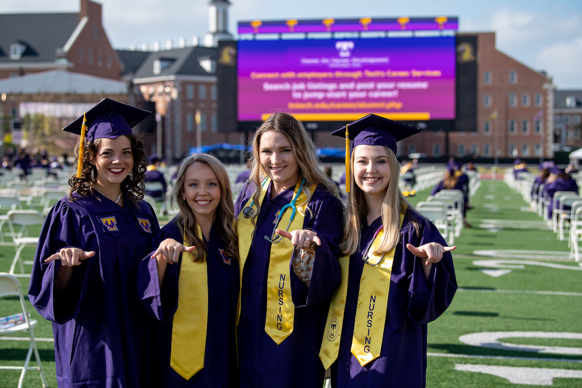 A photo of four nursing students smiling and giving a "wings up" to the camera. They are in purple robes and standing in Tucker Stadium on a sunny day.