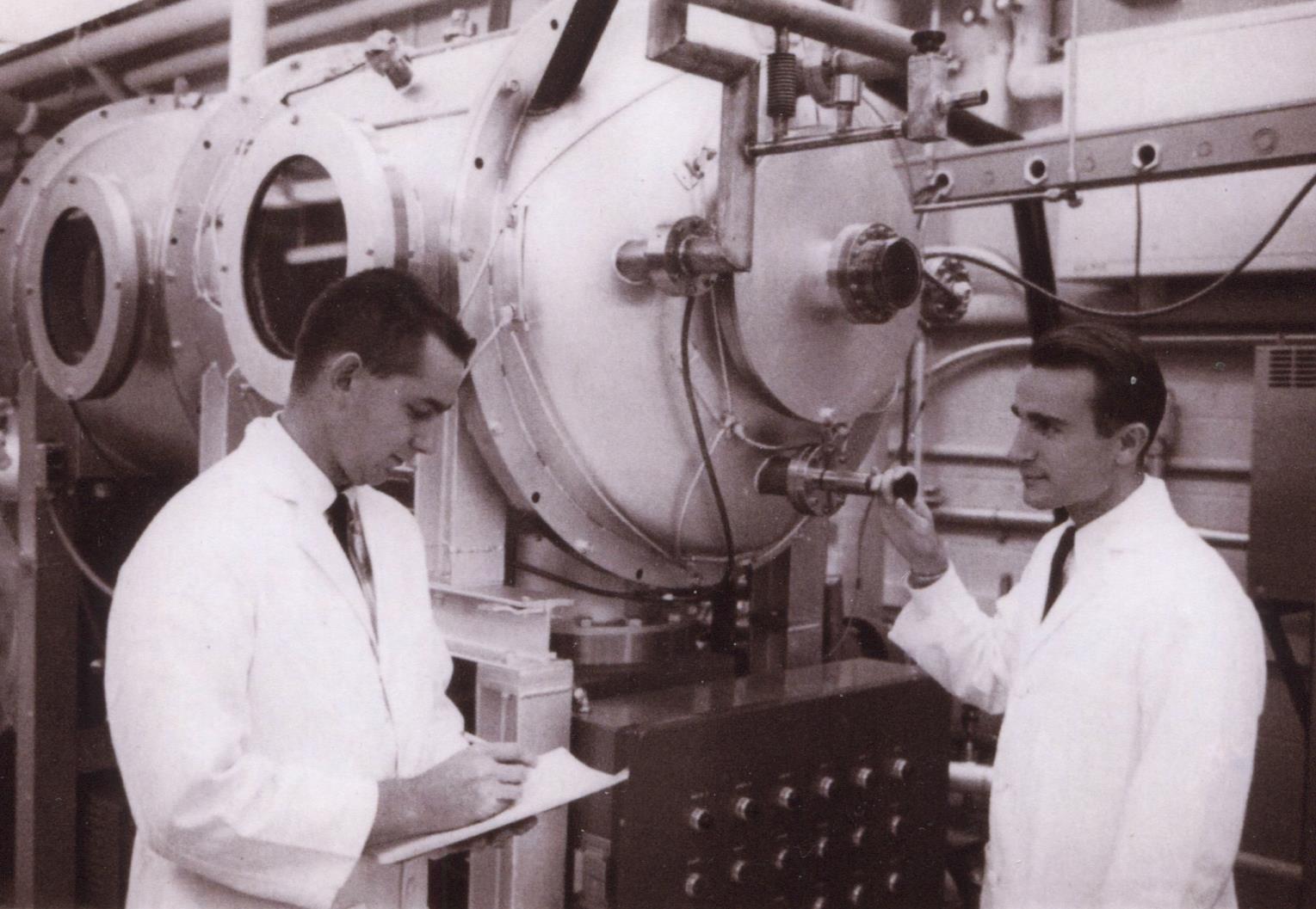 Two gentlemen in white lab coats stand next to a metal capsule with windows in a lab in this 60s era black and white photo.