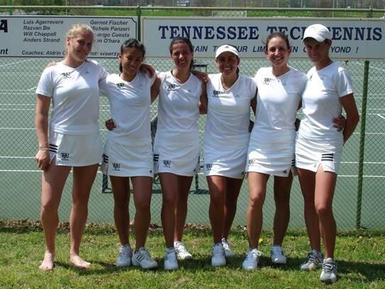 A group of friends dressed in tennis gear stand smiling with their arms around each other in front of a tennis court.