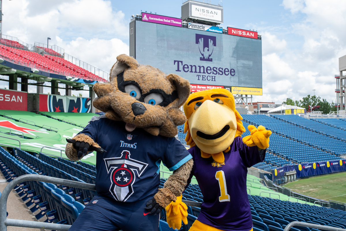 "T-Rac and Awesome Eagle stand in the bleachers at Nissan Stadium on a cloudy day. They are both giving the "wings up" sign. The Tennessee Tech logo is on the jumbotron in the background.