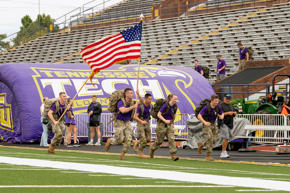 ROTC students running on Overall Field with packs. One student carries the American Flag.