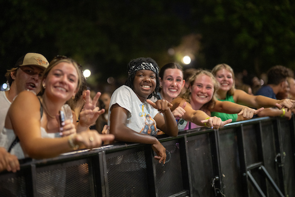 A grould of students leaning on the guardrail at a concert giving a Wings Up to the camera.